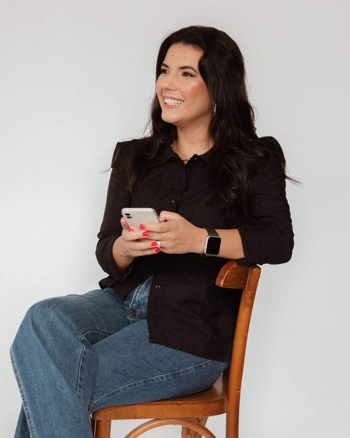 Danieli França sentada, de blusa preta, segurando o celular e sorrindo